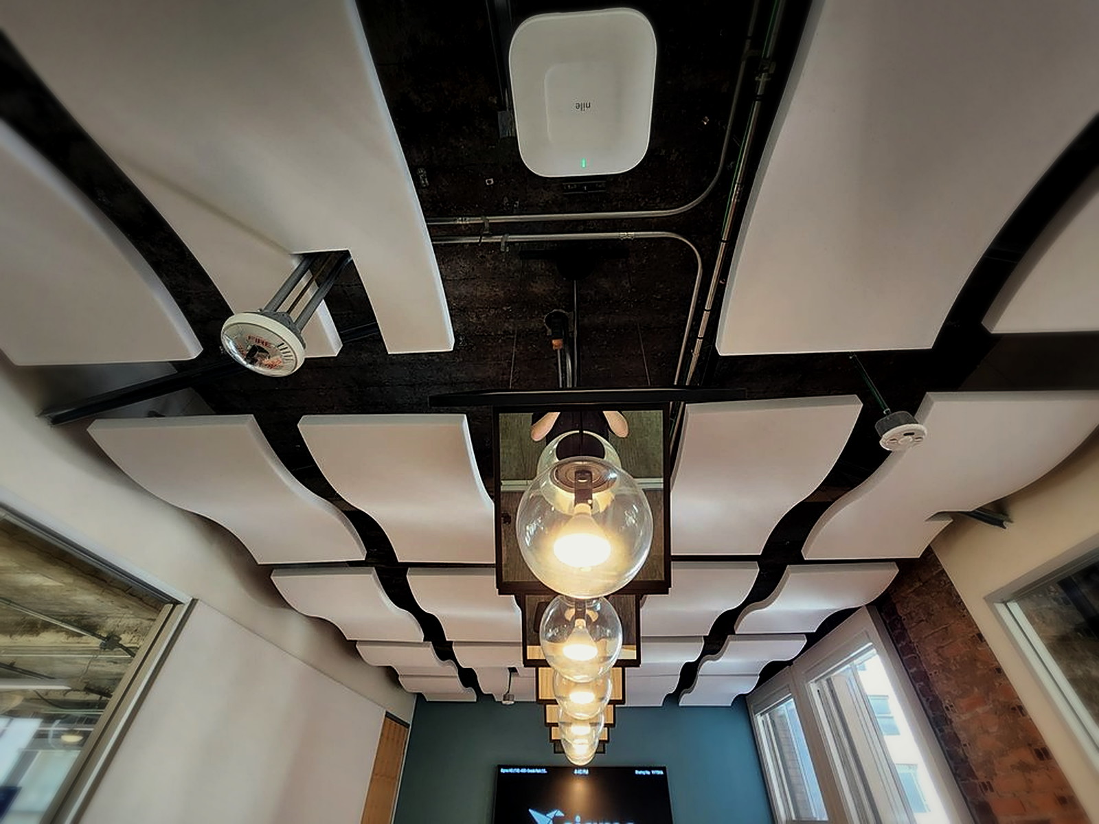 Overhead view of white wave acoustic ceiling clouds installed in a staggered grid under exposed beams and conduit