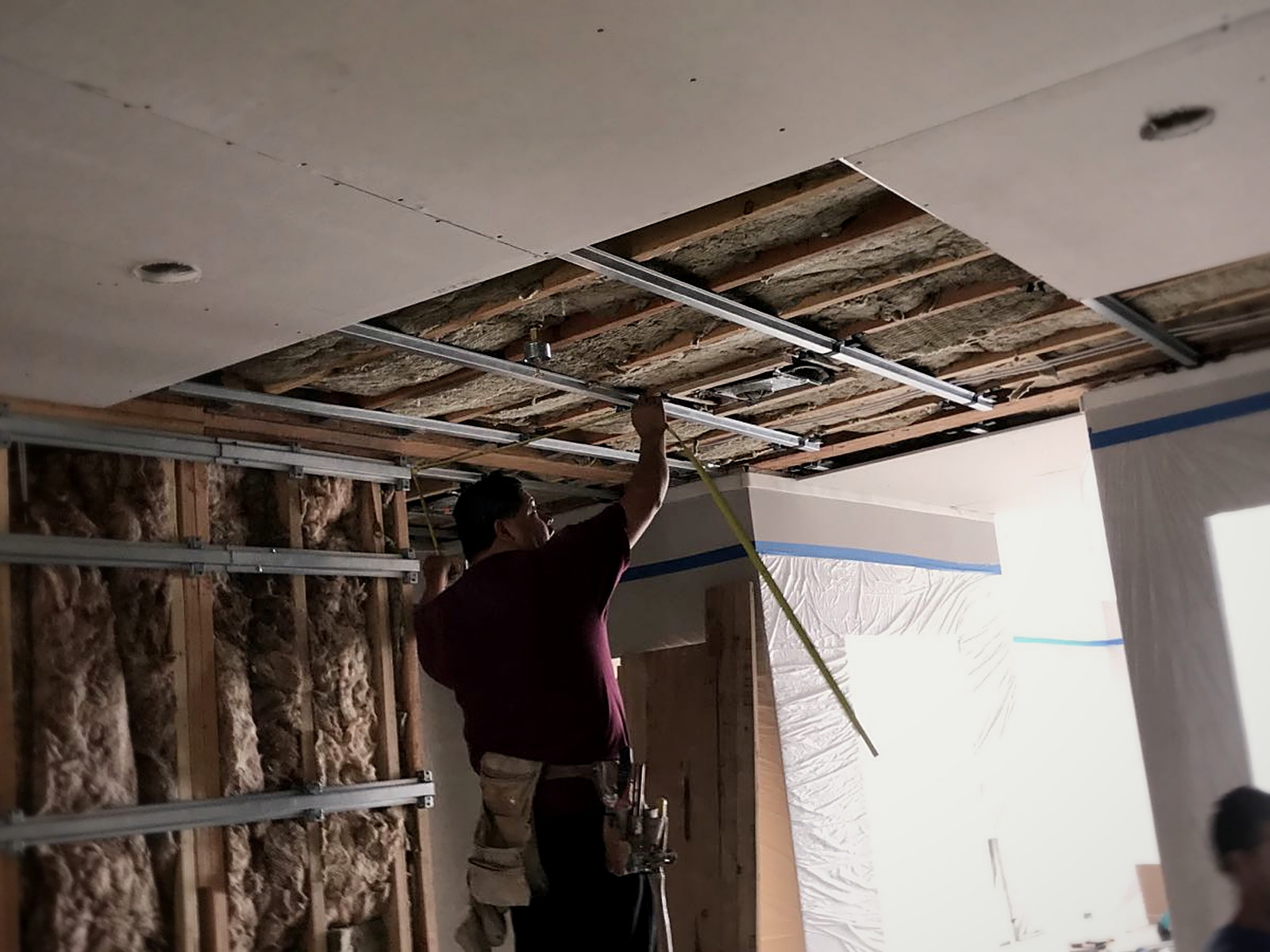 Installer measuring hat channel spacing on a partially drywalled soundproofed residential ceiling