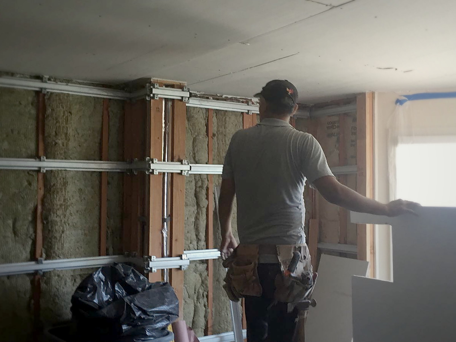 Installer carrying a drywall sheet toward a Melrose wall fitted with insulation, isolation clips and channel
