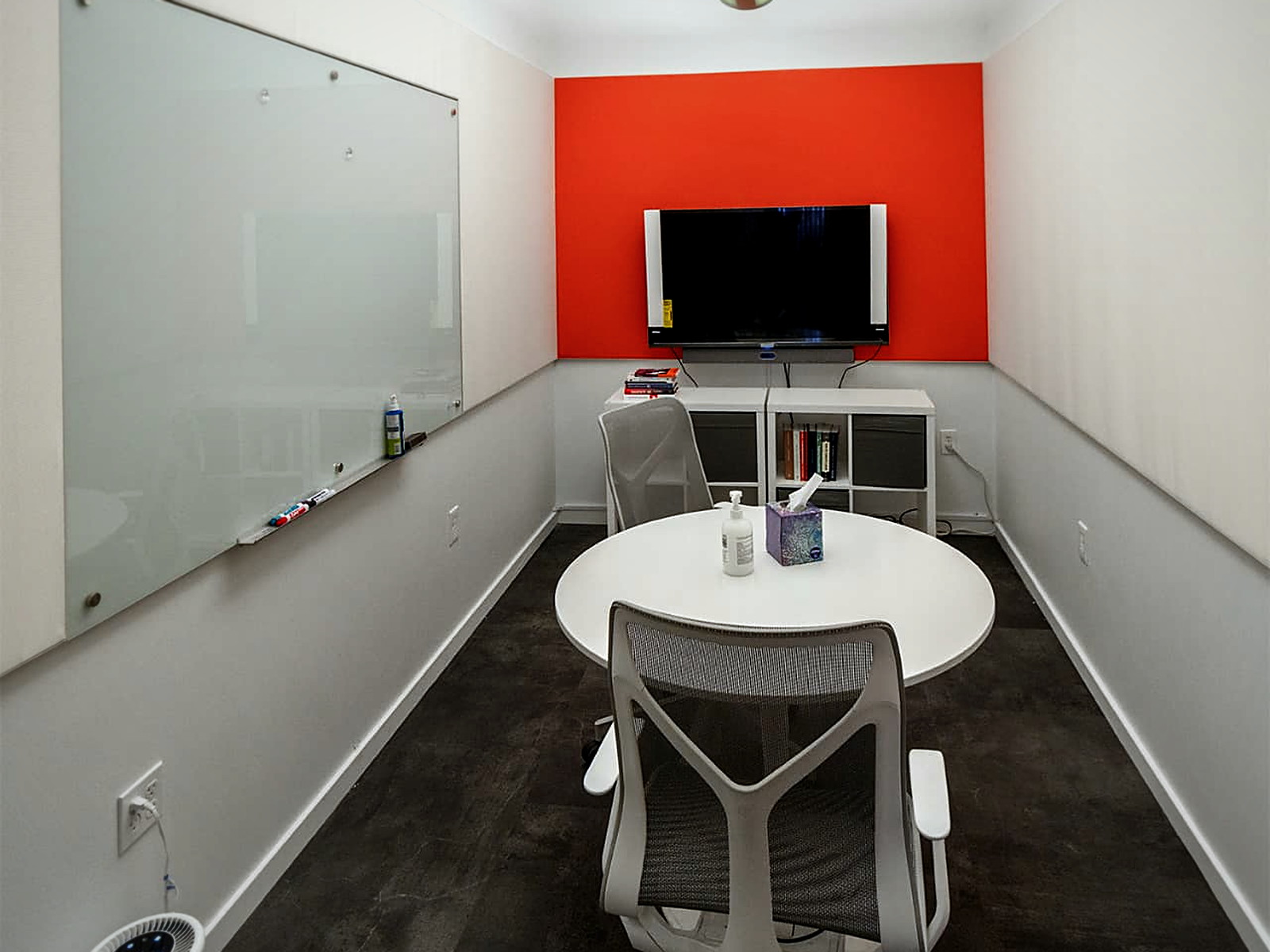 Narrow huddle room with layered acoustic ceiling clouds, a glass whiteboard wall and a display on an orange accent wall