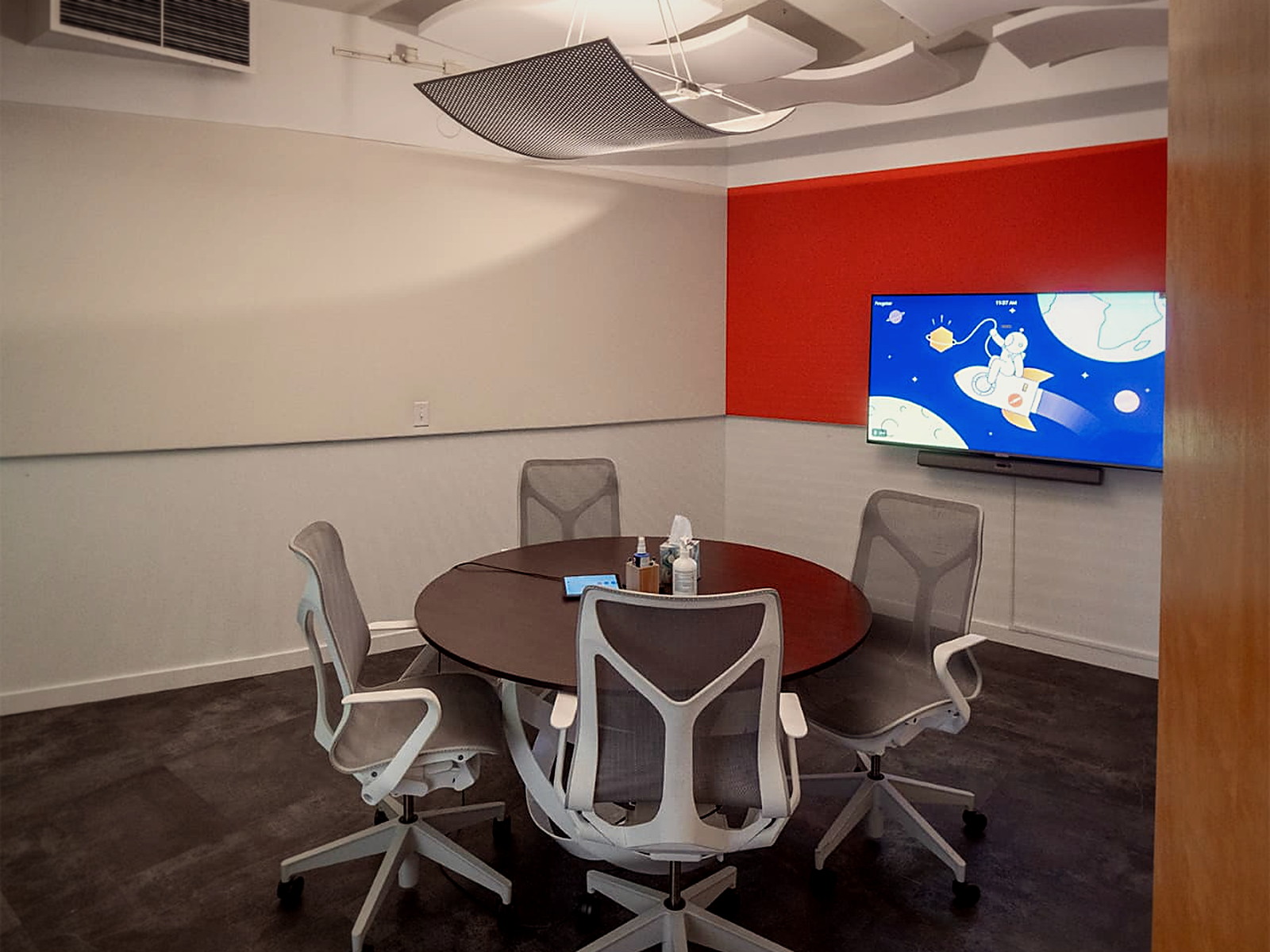 Small round-table meeting room with curved white acoustic ceiling clouds and a wall-mounted display